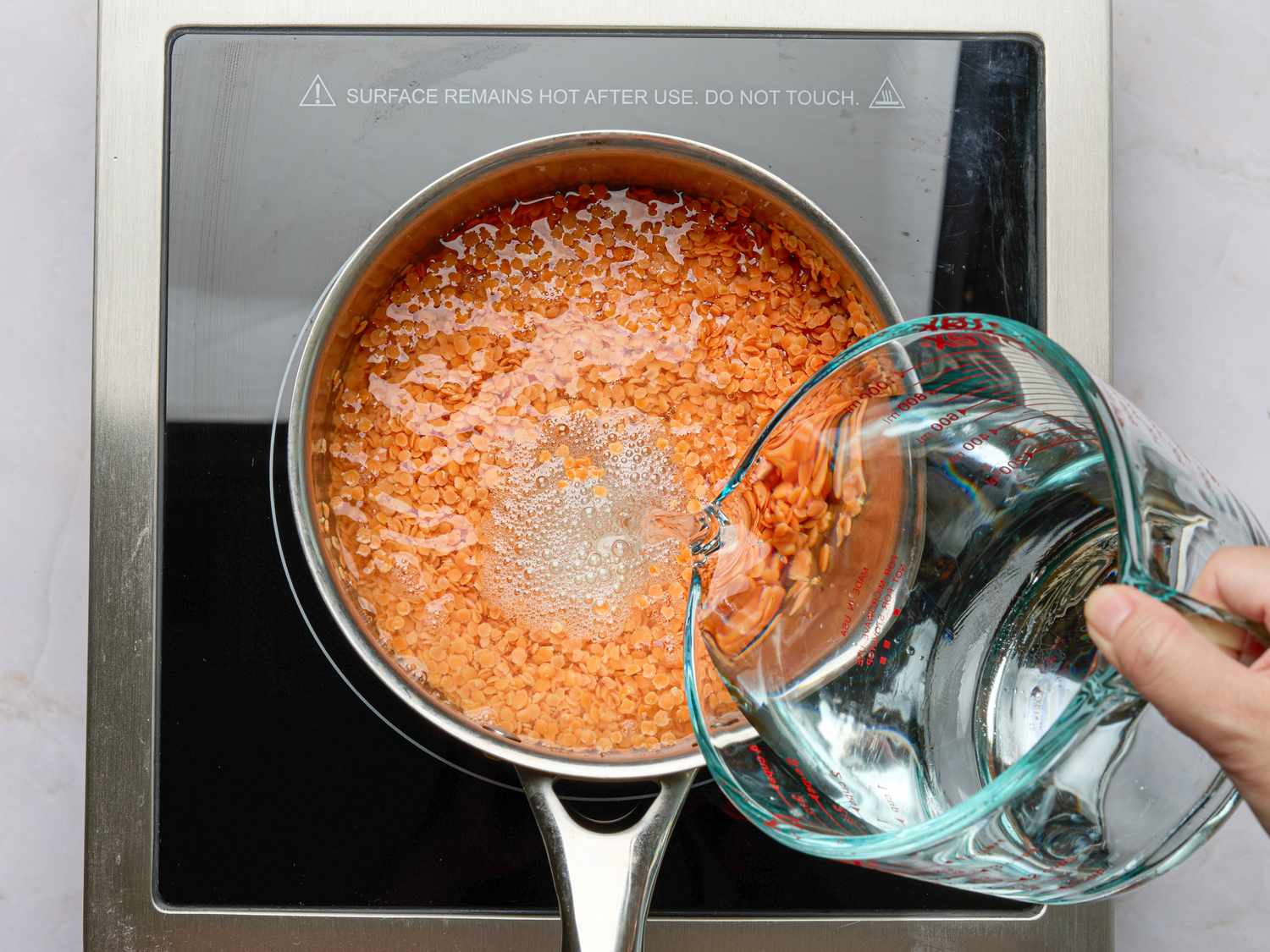 A hand pouring water into a saucepan with ingredients on an induction stove