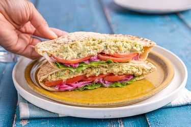 A hand holding a chickpea salad sandwich cut in half, filled with lettuce, red onion, and tomato, placed on a plate