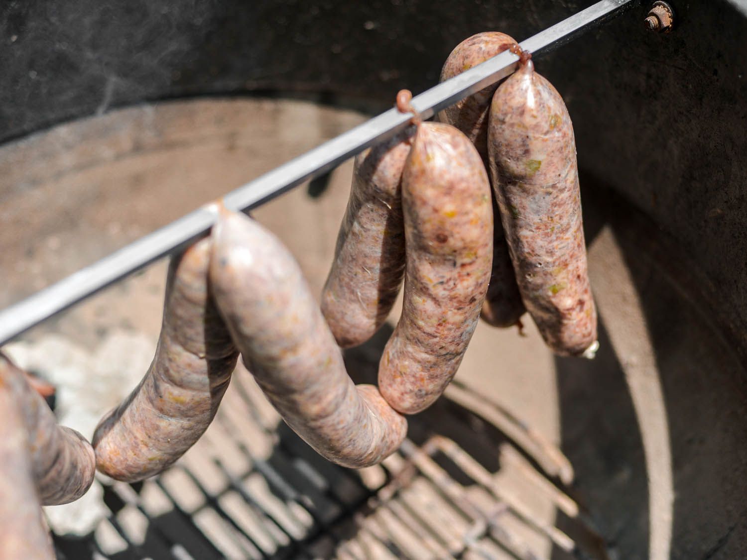 Loukaniko links are hung from a rotisserie spit to cold-smoke in a kettle-style grill.