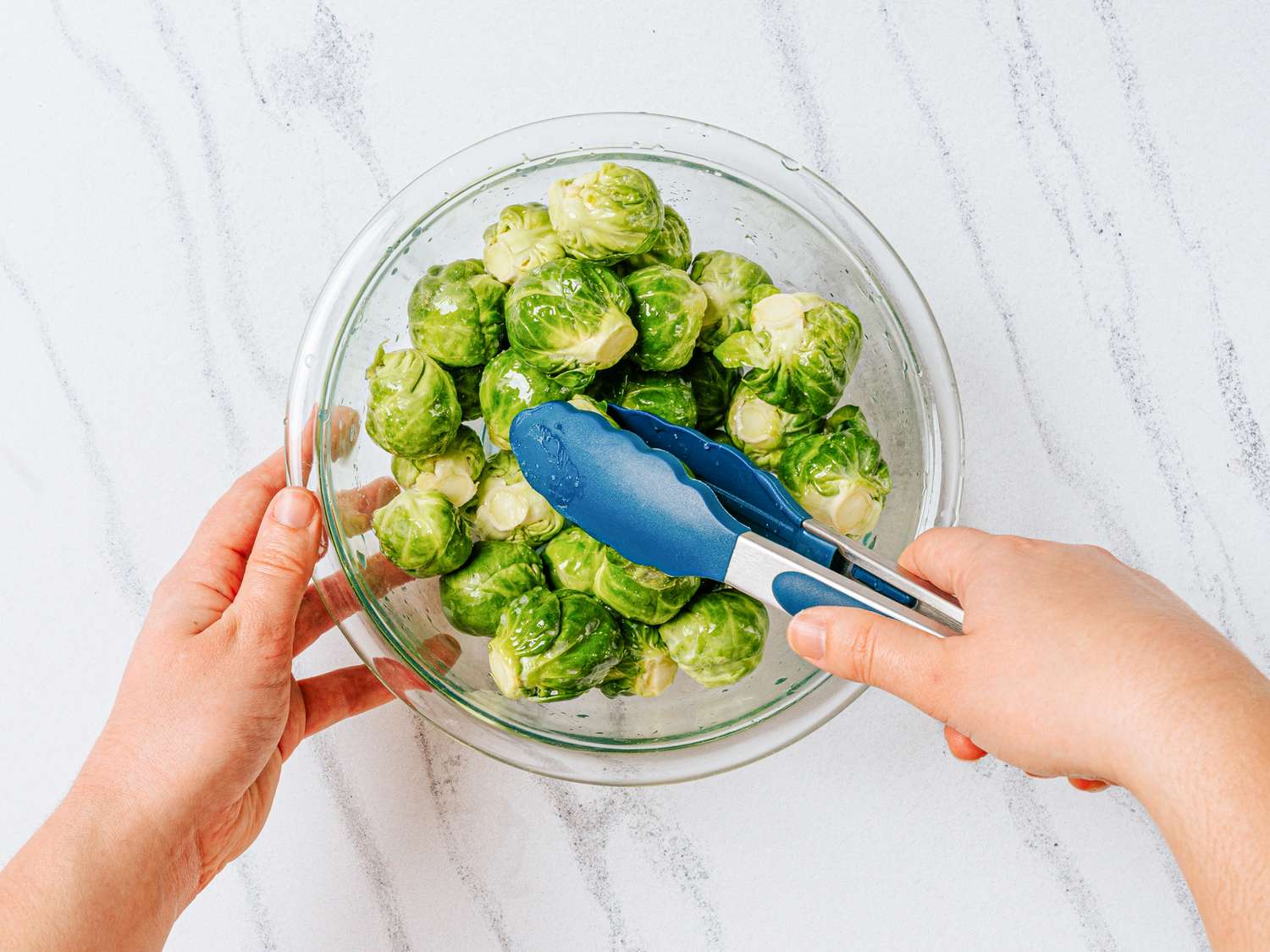 Hands using tongs to toss Brussels sprouts in a glass bowl