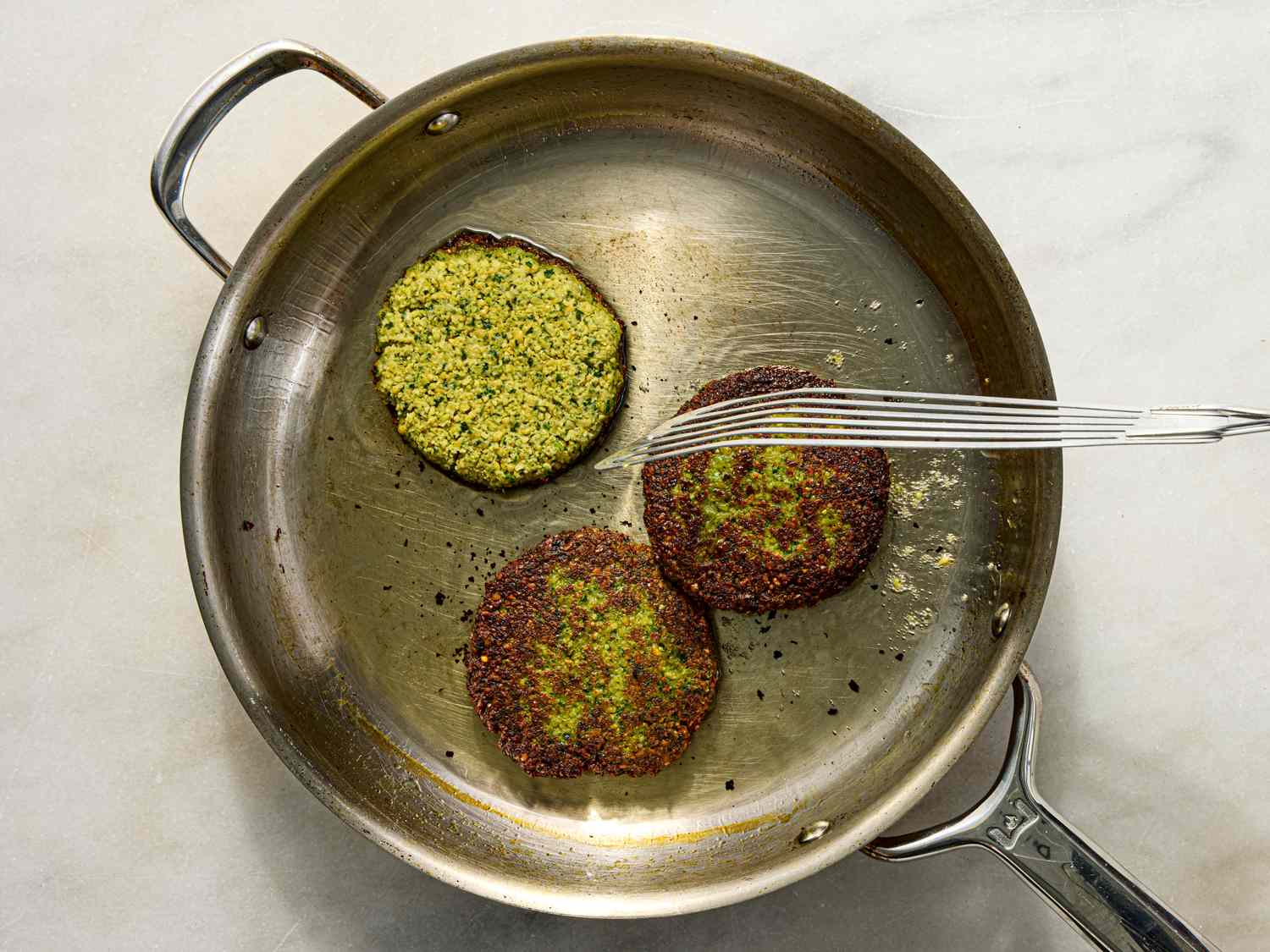A skillet with falafel burger patties being cooked one patty is flipped revealing its texture