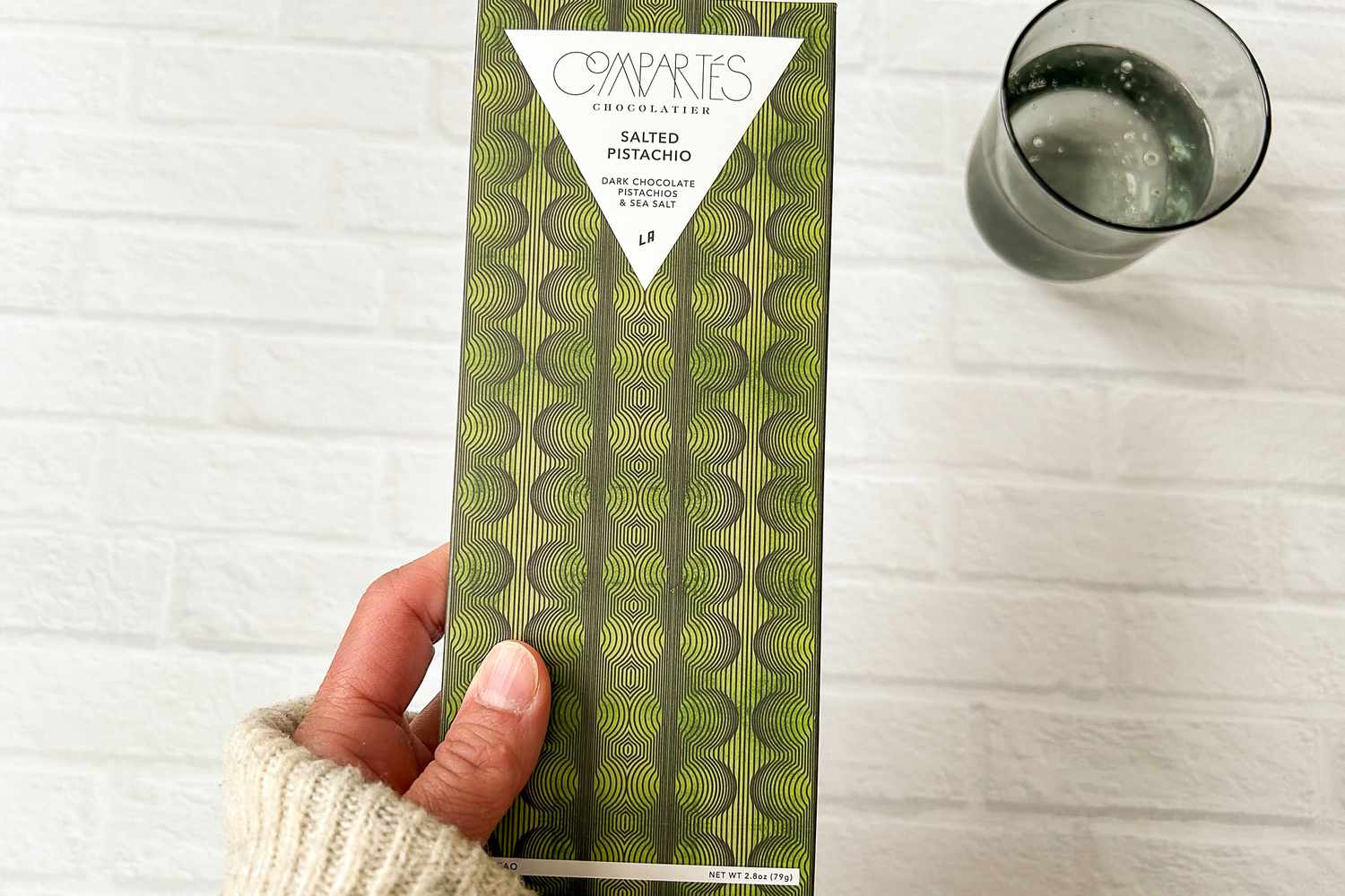 A hand holding Compartés Salted Pistachio Chocolate Bar next to a glass