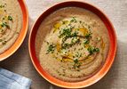 An orange-brown ceramic bowl holding creamy mushroom soup topped with chopped herbs and drizzled olive oil.