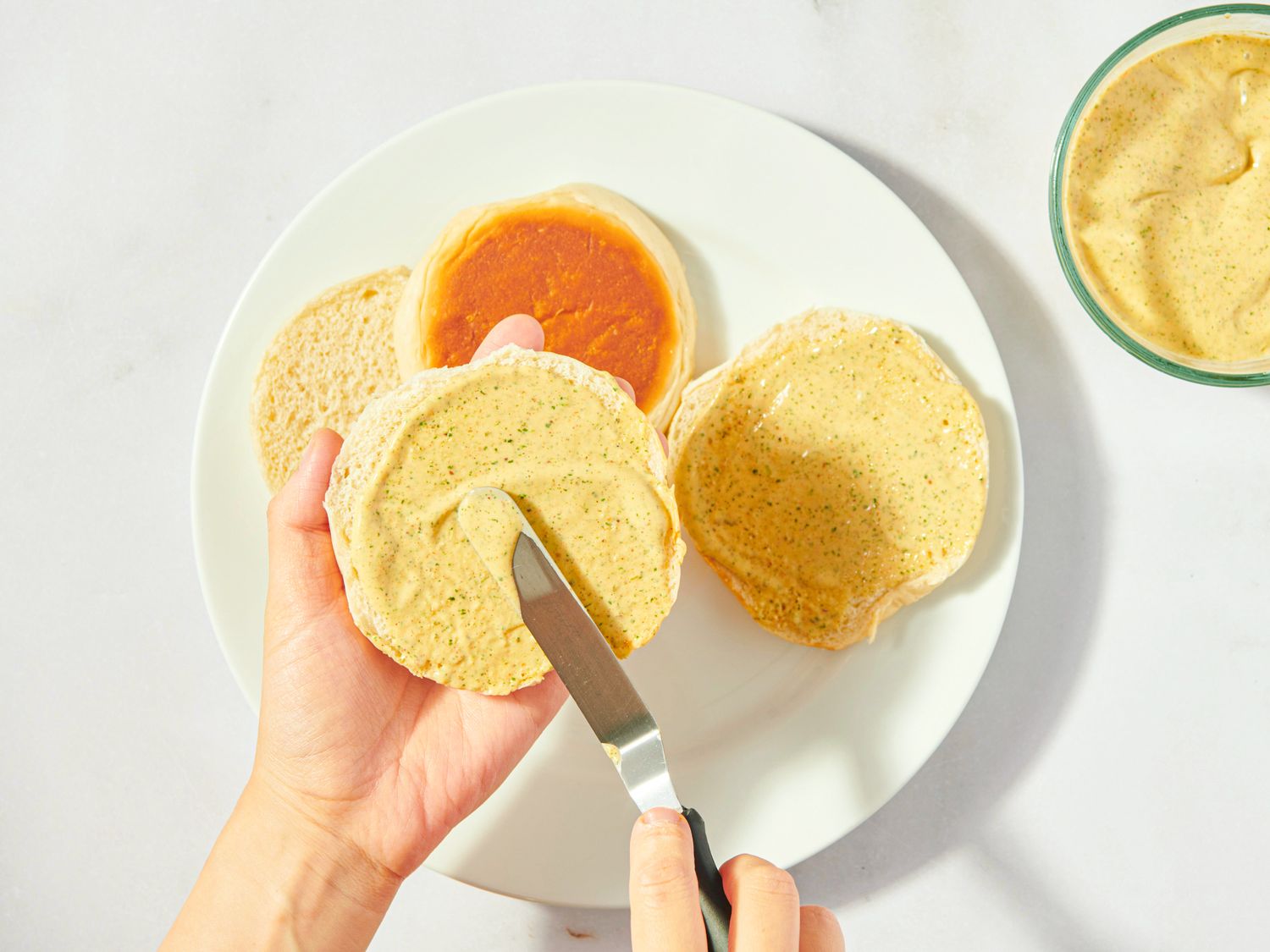 Hands spreading sauce on a sandwich bun next to a plate with other prepared buns and sauce