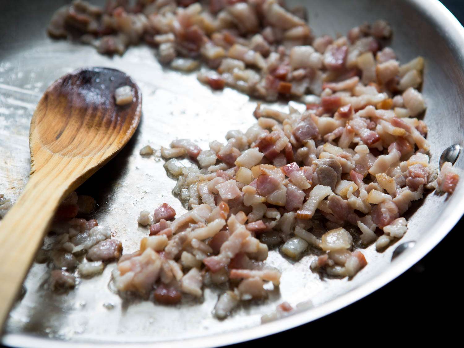 Diced bacon being browned in a stainless steel skillet with a wooden spoon on the left side of the frame