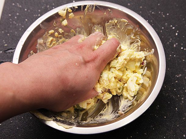 Author's hand reaching into a bowl of cooked eggs, squeezing and mashing them to a chunky-yet-creamy texture.