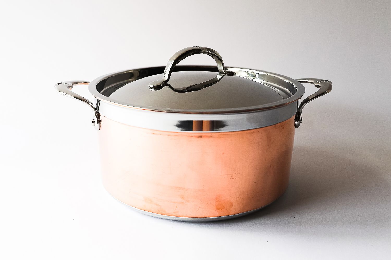 A polished metal pot with a lid and handles on a neutral background