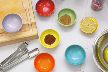 An assortment of small colorful bowls containing spices next to kitchen tools wooden board and a lemon half
