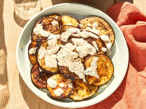 Grilled eggplant slices topped with a creamy white sauce served in a bowl on a wooden table beside a red cloth napkin