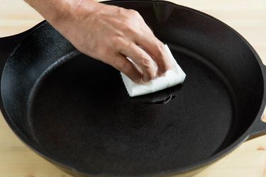 A hand holding a folded paper towel, applying oil to the inside of a large cast iron pan.