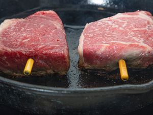 Two pieces of steak being cooked in a castiron skillet with yellow thermometers inserted