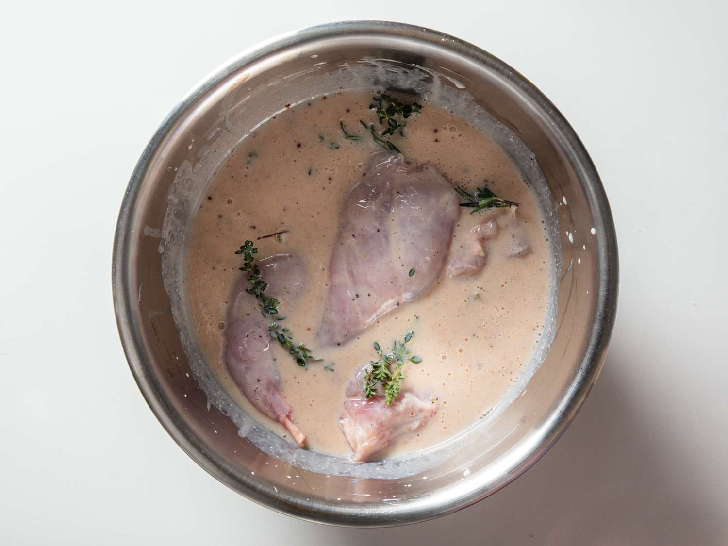 Rabbit pieces submerged in bowl of buttermilk brine with herb sprigs