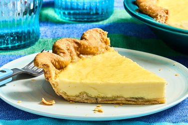 Slice of custard pie on a white plate with a fork next to it, surrounded by blue glasses and a colorful tablecloth