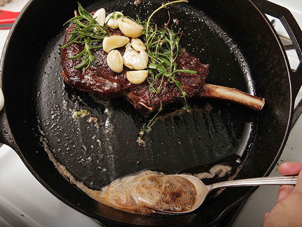 Tilting skillet to pull basting butter away from browned steak, garlic cloves, and herbs to spoon