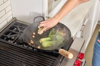 A person cooks bok choi in the Mammafong Pre-Seasoned Blue Carbon Steel Flat-Bottom Pow Wok
