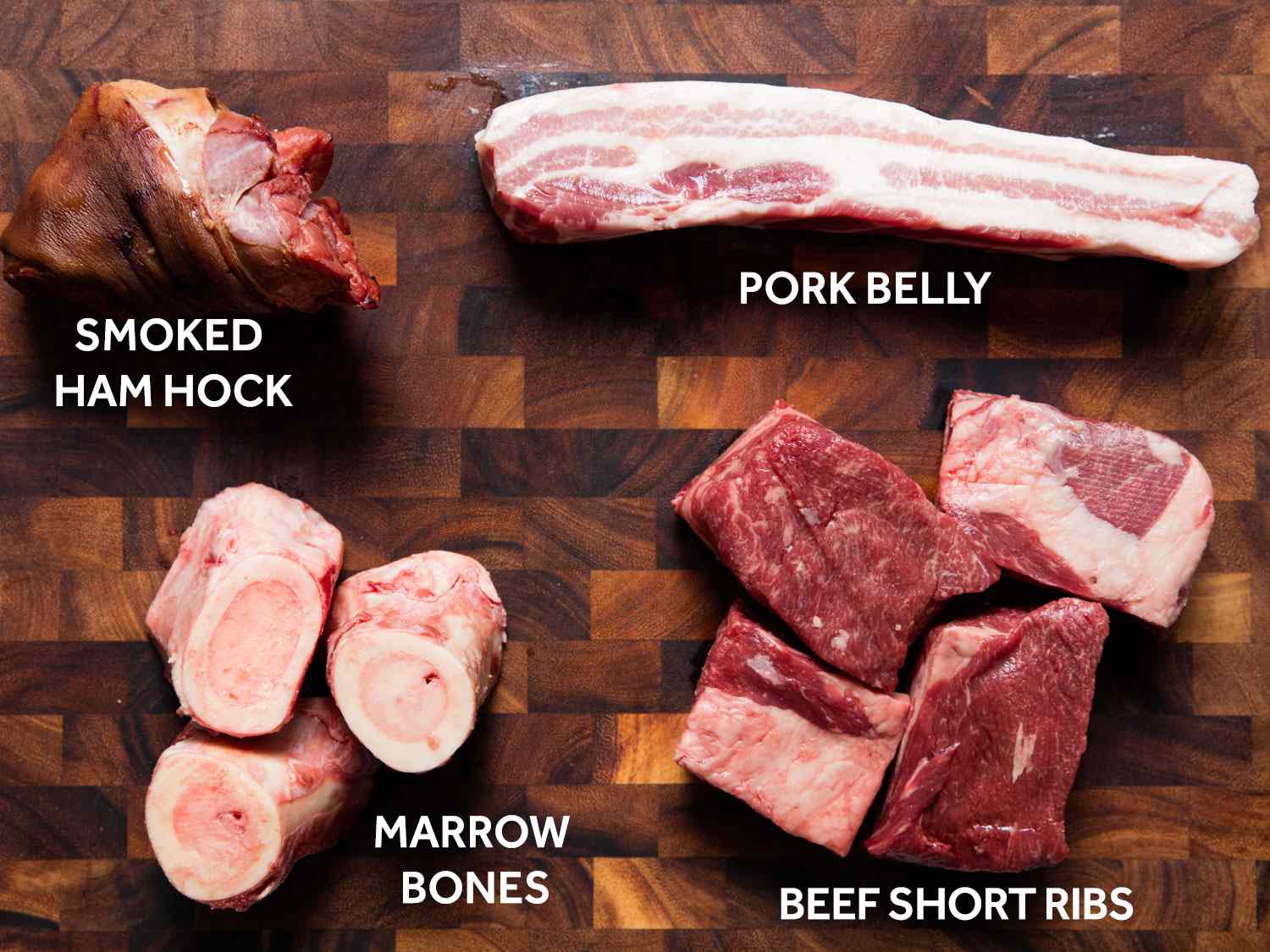 Overhead shot of raw pork belly, beef short ribs, marrow bones, and smoked ham hock on a wooden surface.