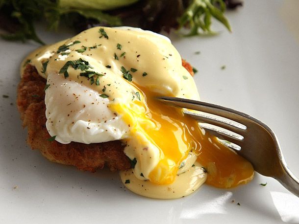 A poached egg with running yolk resting on a crab cake on a white plate. 