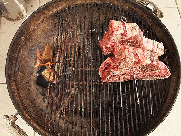 The two skewered porterhouses standing upright on a grill grate, opposite a small charcoal fire with two pieces of hardwood placed on it.