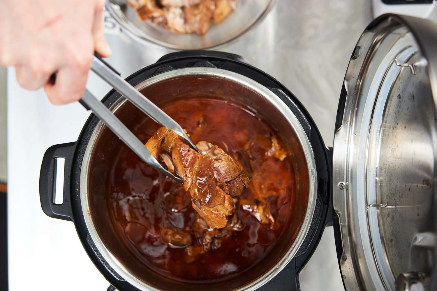 A person using tongs to remove cooked meat from a pressure cooker filled with sauce seen on a kitchen counter