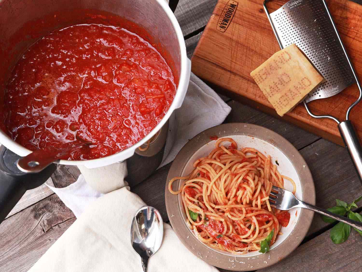 Overhead shot of a pot of pressure cooker tomato sauce, bowl of pasta tossed in tomato sauce, and hunk of parmigiano resting on a cheese grater