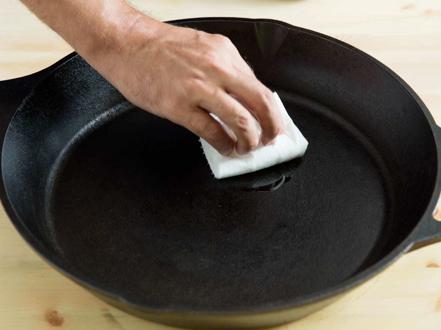 A hand holding a folded paper towel, applying oil to the inside of a large cast iron pan.