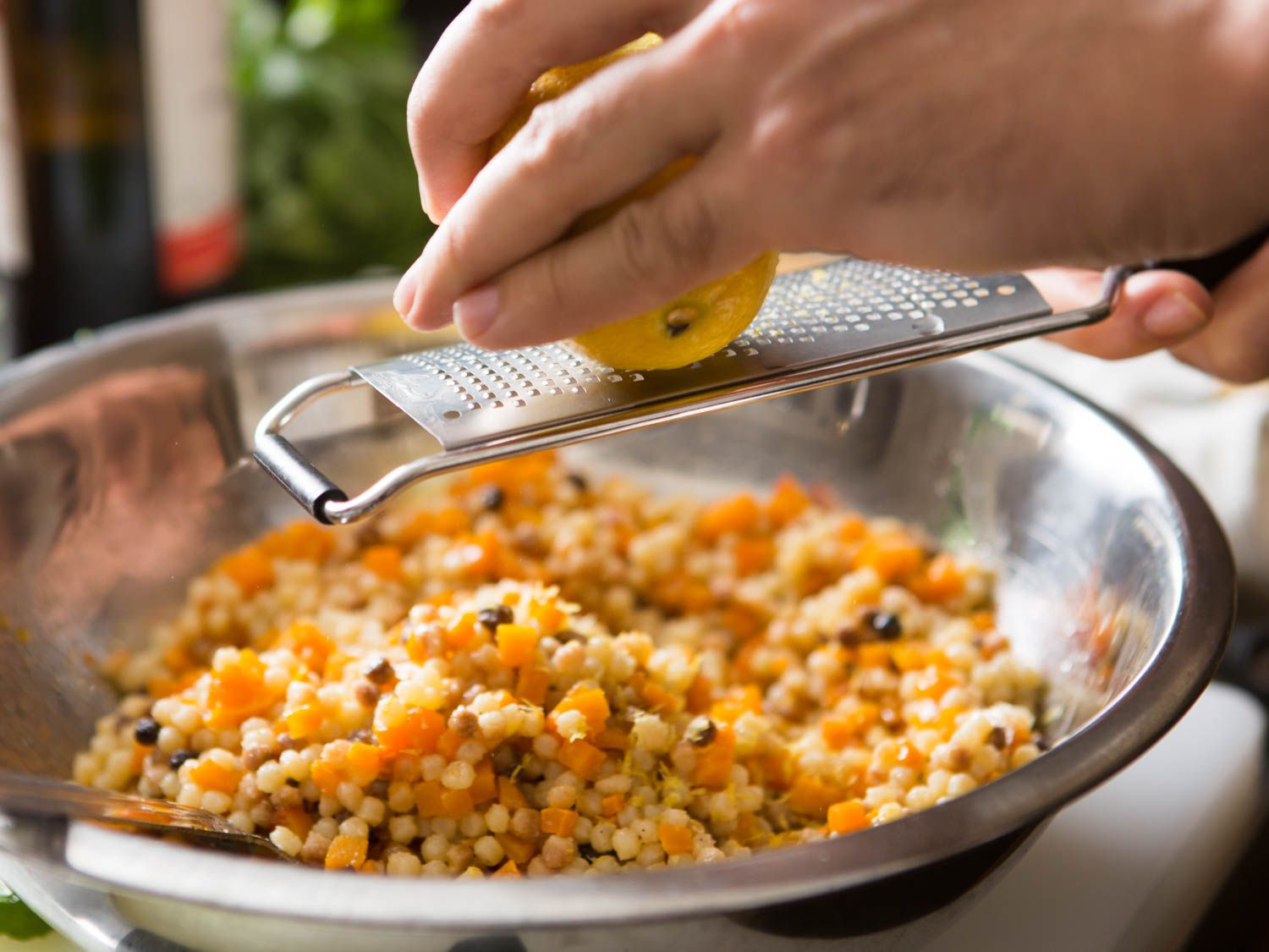Zesting a lemon with a grater over a bowl of fregola and squash.