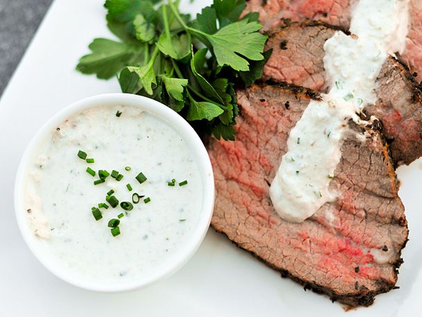 A small ramekin of horseradish sauce on a plate of sliced roast beef with horseradish cream spooned over top. 