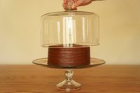 A glass cake stand holds a chocolate cake; a hand lifts a glass dome off the stand.