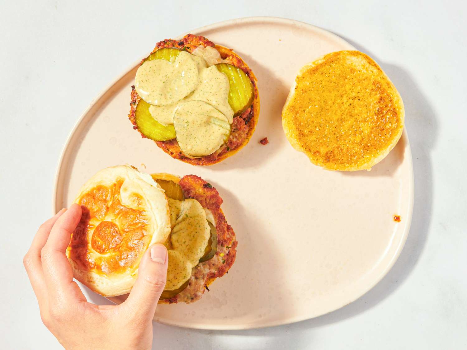 A smash burger being assembled with pickles and cheese on a plate hand placing top bun