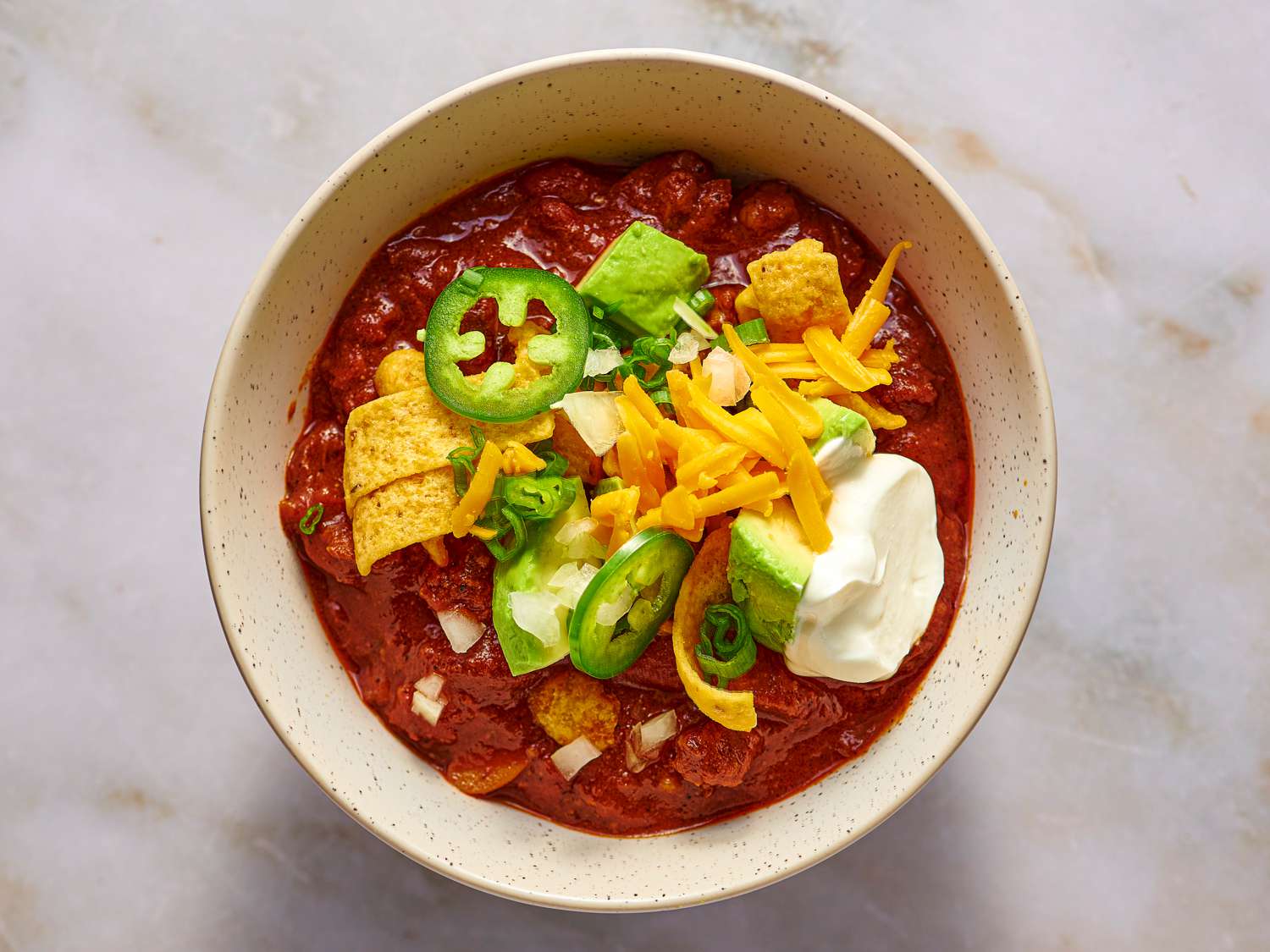 A ceramic bowl of chili topped with sliced jalapenos, sliced scallions, grated cheddar, sour cream, and corn chips.
