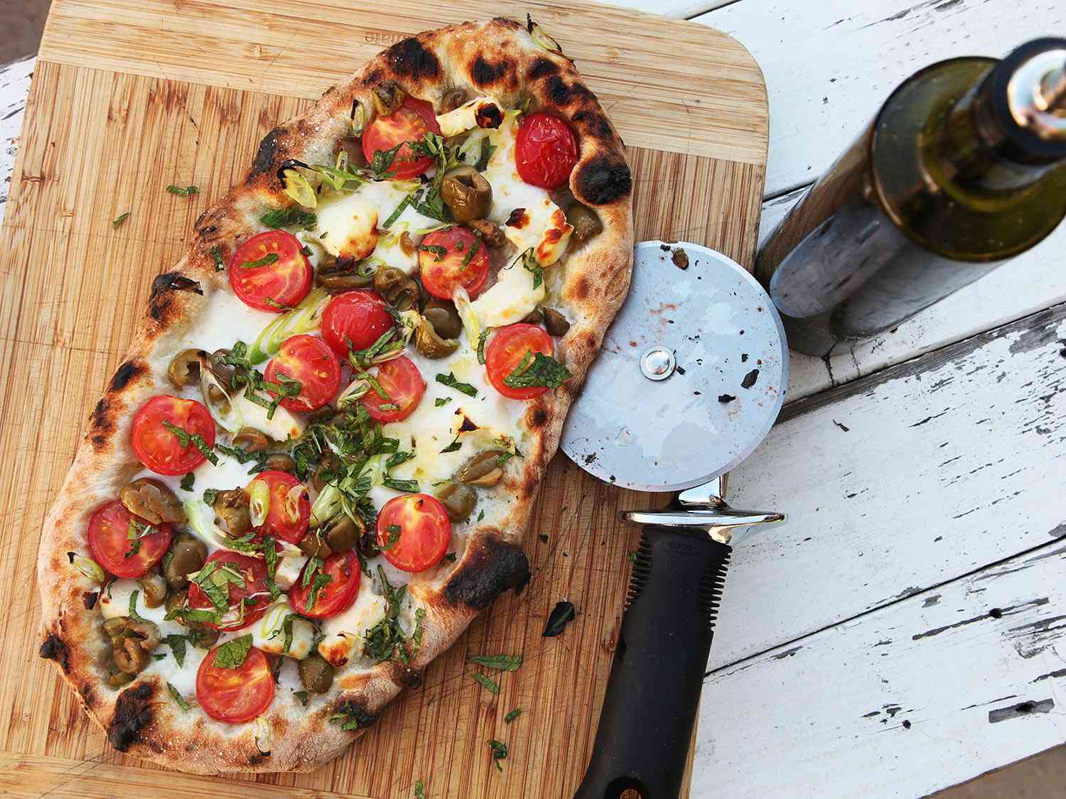 A halloumi, cherry tomato, and olive flatbread on a cutting board outside.