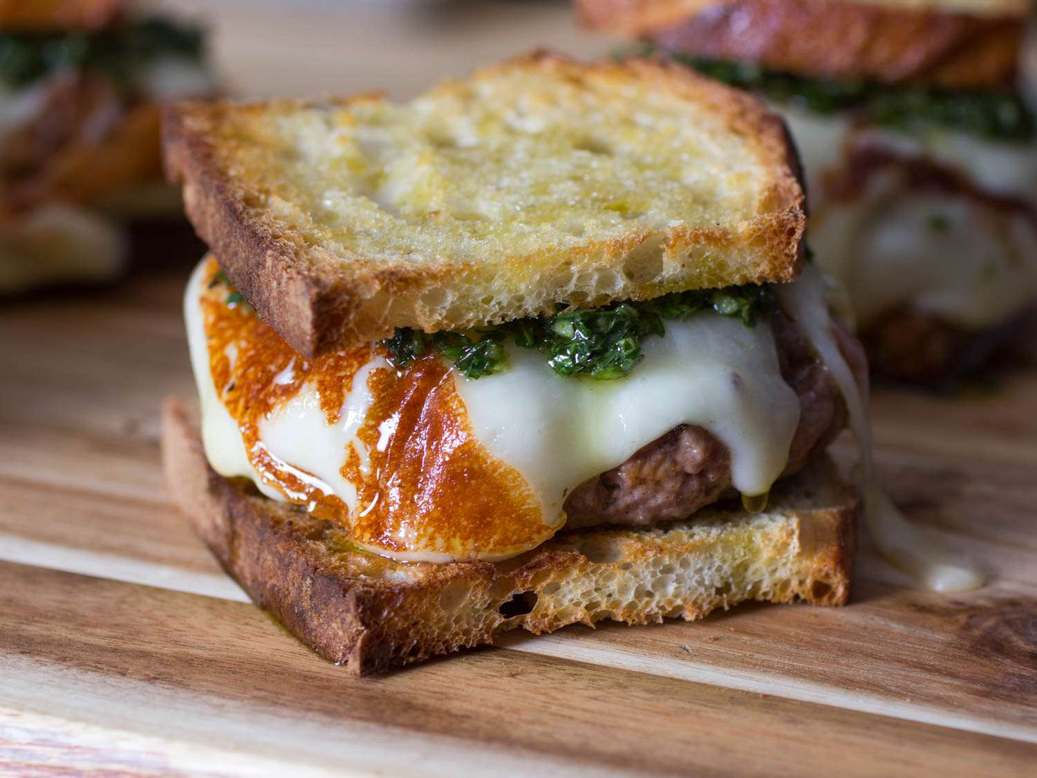 Three asado burgers on a wood cutting board, each made on sliced bread and topped with melted cheese and chimichurri. 