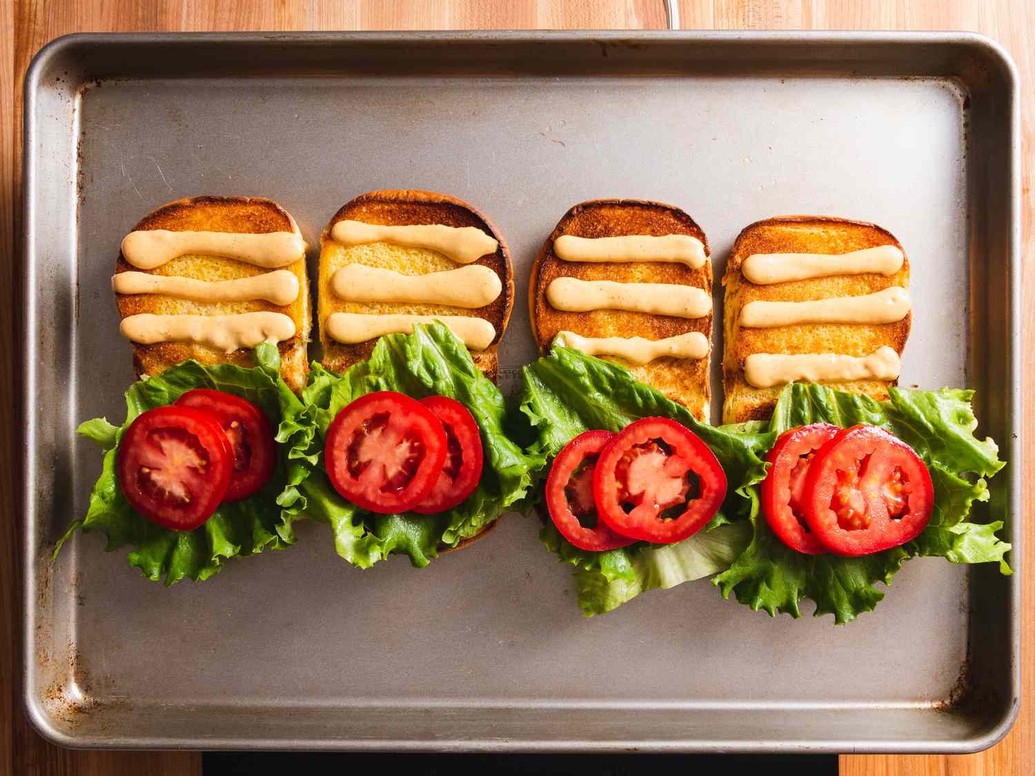 Four toasted burger buns open on a baking sheet, each one with three lines of homemade Shack Sauce on the top half of the bun, and lettuce and tomato on the bottom half of the bun.