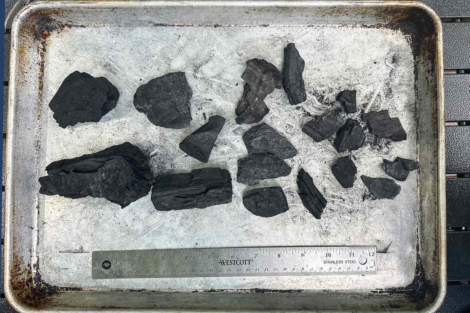 A sheet pan with assorted sizes of lump charcoal pieces on it. A ruler beneath shows the sizes.