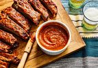overhead view of bbq sauce on a cutting board alongside ribs and glasses of beer