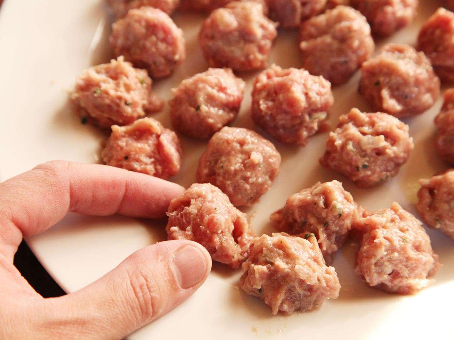 A hand picking up one of several very small uncooked meatballs.