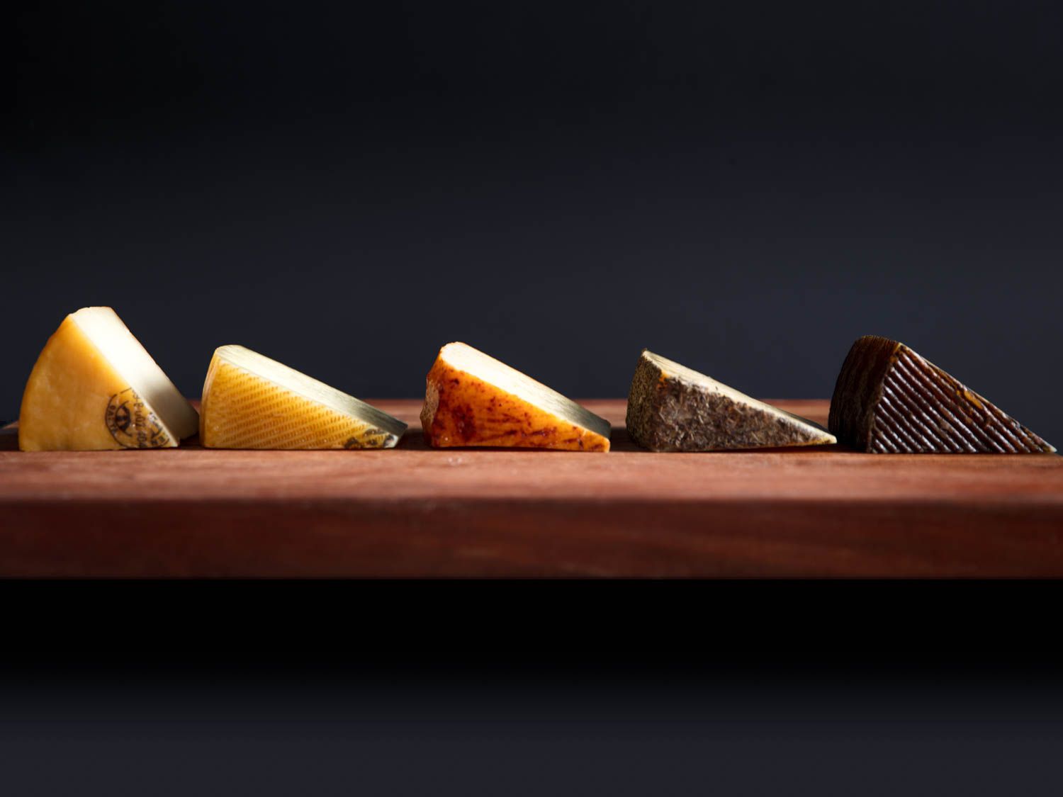 Profile view of five different Manchego-type cheeses, cut into wedges and arranged on a wooden board.