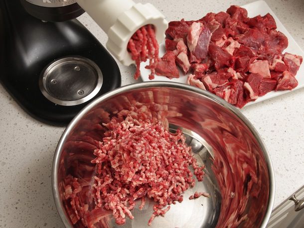 Grinding beef with a stand mixer fitted with a grinding attachment. Chunks of beef are piled on a platter, waiting to be fed into the grinder's chute. Ground meat is being extruded from the grinder plate and falling into a mixing bowl. 