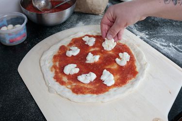 A hand laying mozzarella on a pizza atop a wooden pizza peel