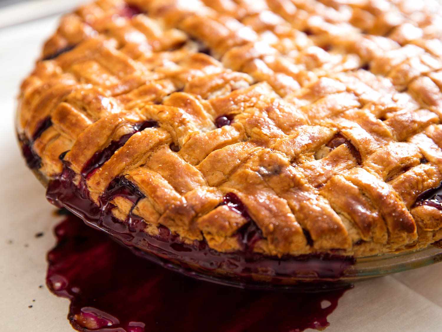 Close-up of the summer fruit pie, fresh from the oven. The basketweave crust is golden brown and some of the filling's juices have bubbled out onto the parchment-lined rimmed baking sheet underneath.