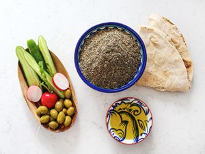 Small bowl of Za'tar next to a piece of pita, a small bowl of olive oil, and a rectangular bowl of vegetables