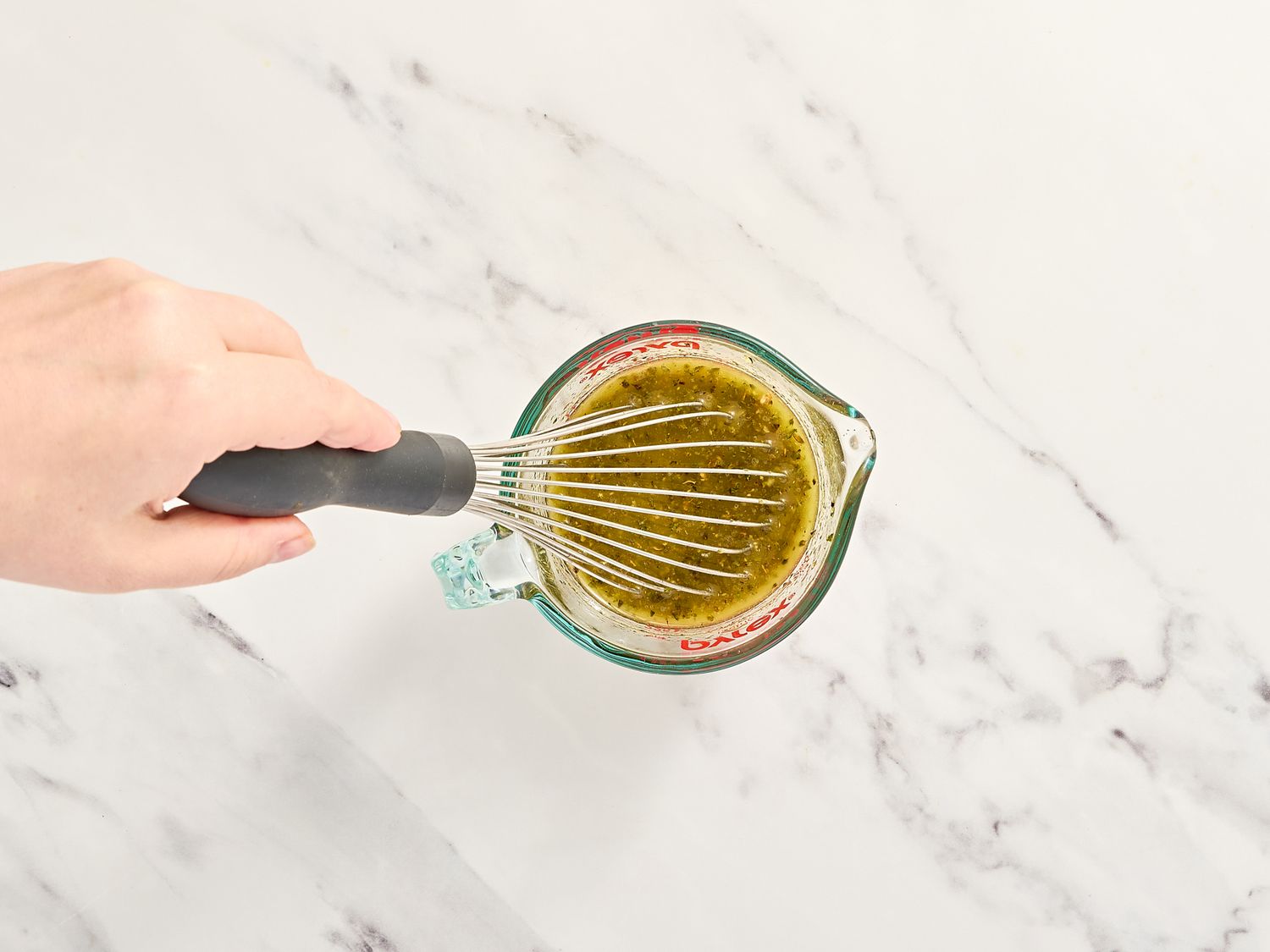 A hand whisking a sauce in a glass measuring cup on a white marble surface