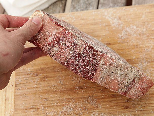 Author's hand lifting one side of a seasoned porterhouse to show the application of salt and pepper to the side of the steak.