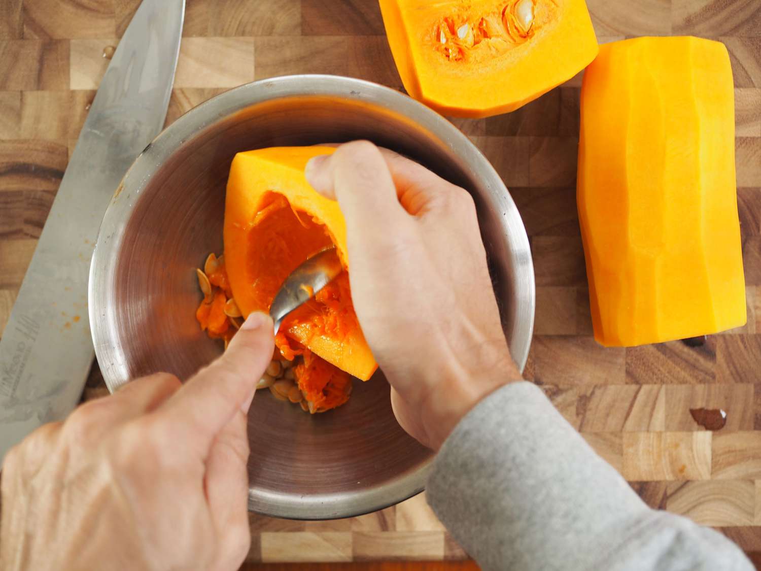 A pair of hands over a bowl, with one hand scraping out the cavity of a butternut squash, and the other holding the squash half.