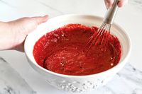 A hand stirring a red batter in a Mason Cash bowl with a wire whisk