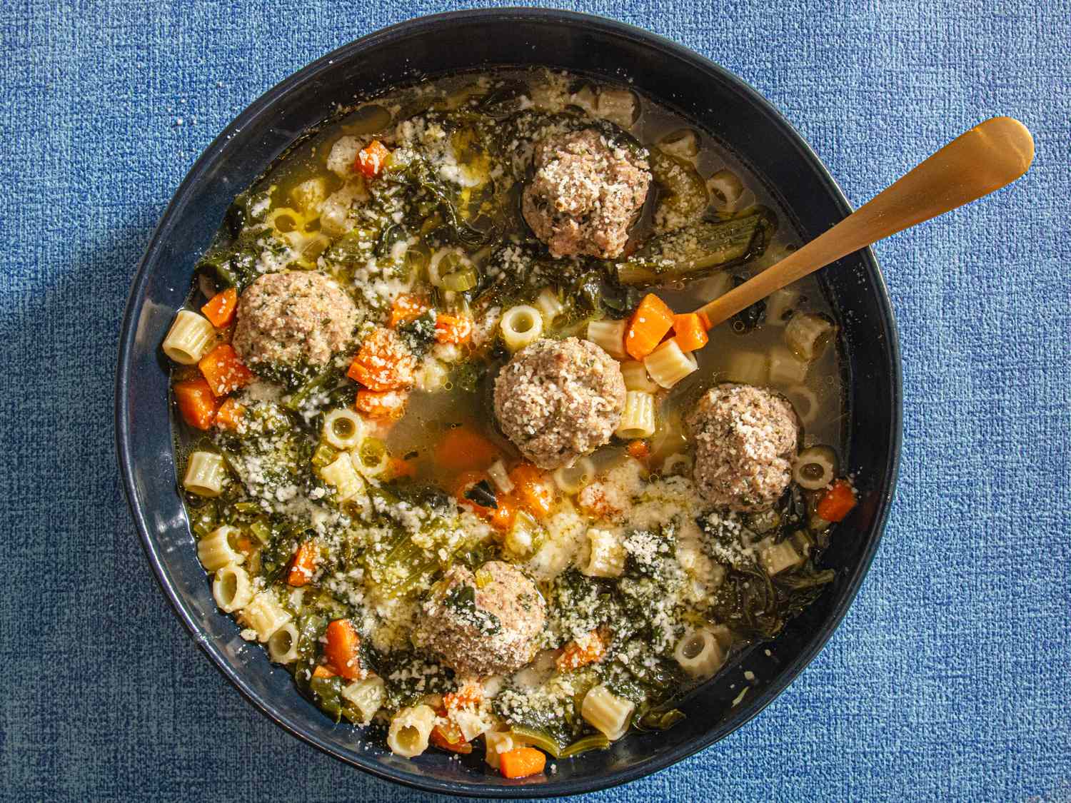 A bowl of Italian wedding soup with meatballs pasta and vegetables served on a blue surface
