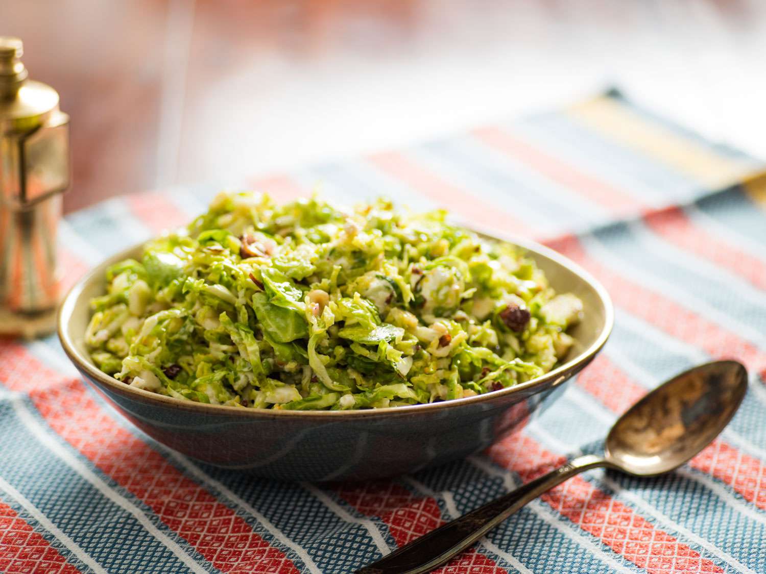 Salt-wilted Brussels sprout salad with hazelnuts and goat cheese in a bowl next to a spoon and a pepper grinder on a striped napkin.