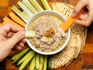 Two hands holding slices of vegetable dipping into a bowl of walnut skordalia surrounded by flatbread and vegetable sticks