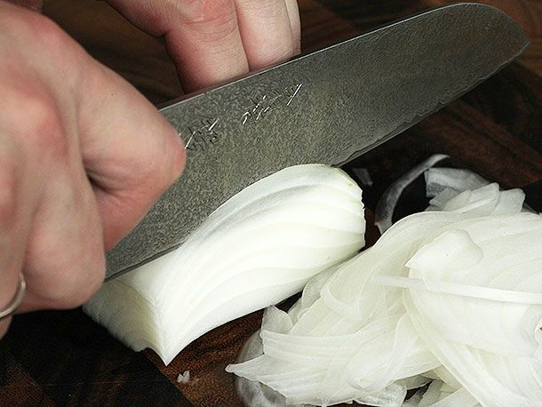A knife held parallel to a cutting board slicing straight down into half of an onion.