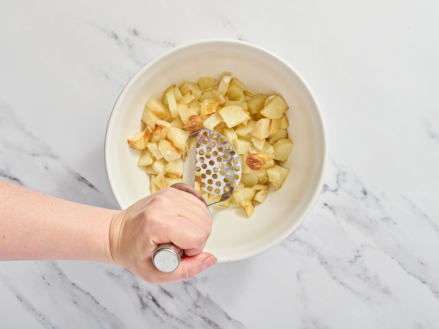 A hand mashing cooked cubed potatoes in a bowl with a masher